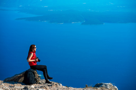 Young woman photographer sitting on the top of mountain with photo camera on blue sea backgroundの写真素材