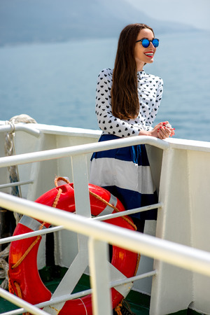 Young woman dressed in shirt and skirt sailing on cruise liner or ferry with sea and mountain backgroundの写真素材