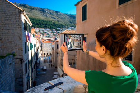 Young woman photographing with digital gadget old buildings in Dubrovnik old city center. Back view with tablet screen.の写真素材