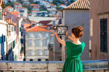 Young woman photographing with digital gadget old buildings in Dubrovnik old city center. Back view with tablet screen.の写真素材