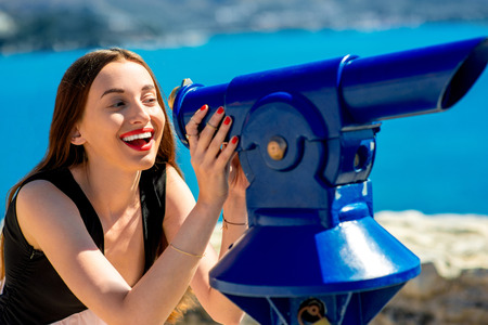 Young woman traveler looking through a blue telescope outdoors near the seaの写真素材