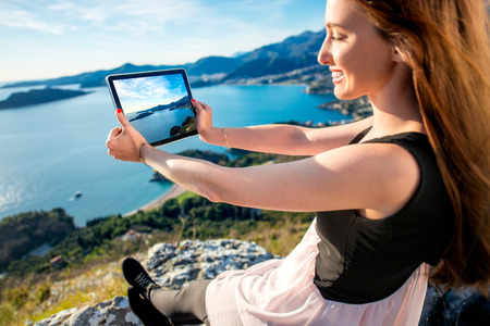 Young woman in dress photographing with digital table sea scape sitting on the mountain topの写真素材