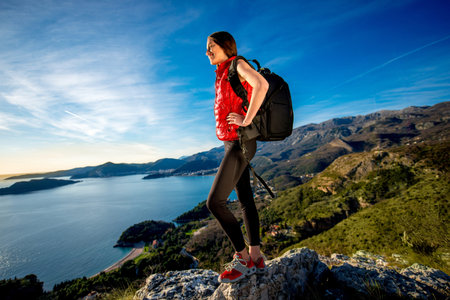 Sports woman in red vest with backpack standing on the top of mountain on the sunset.