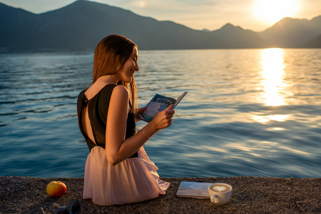 Young woman reading book and looking at beautiful sunrise on the pier with sea and mountains on backgroundの写真素材