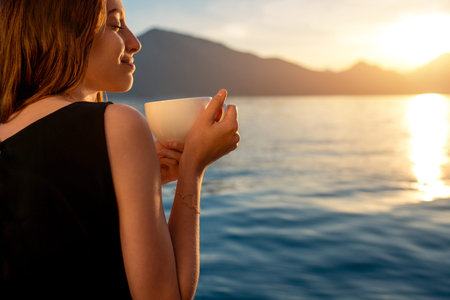 Young woman enjoying coffee on the pier at sunrise with mountains on backgroundの写真素材