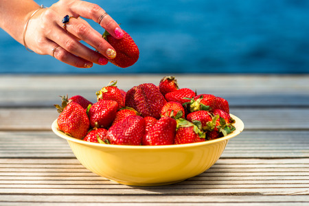 Taking with hand fresh strawberry from the yellow plate outdoors on the blue backgroundの写真素材