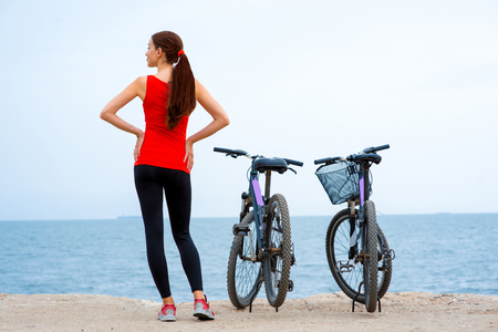 Sport woman in red shirt standing with bicycles on the concrete seacoastの写真素材