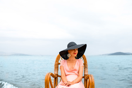 Young woman in pink dress and big black hat resting in the wicker chair on the beach in cloudy weather.の写真素材