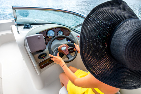 Young and pretty woman in yellow skirt and swimsuit with hat and sunglasses driving luxury yacht in the sea. Top view without face focused on hands and helmの写真素材