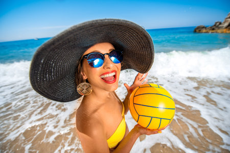 Playful woman in big hat with the yellow ball standing on the beach.