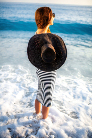 Young and elegant woman in stripped dress with a hat standing on the beach on sunsetの写真素材
