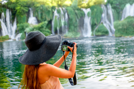 Young woman tourist in pink dress and big hat photographing with professional photo camera beautiful Kravica waterfall in Bosnia and Herzegovinaの写真素材