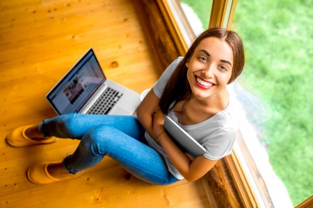 Young and cute woman in jeans, slippers and white shirt sitting on the floor holding digital tablet near the window in cozy wooden cottageの写真素材