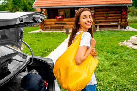 Young and attractive woman with yellow back standing near the car with wooden house on background. Countryside vacations conceptの写真素材