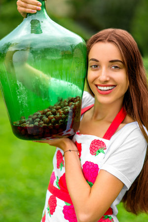 Young and healthy woman in apron holding big green bottle with red berries in the garden
