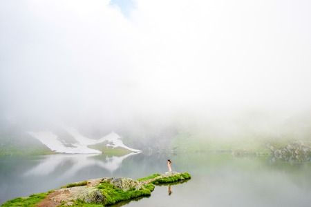 Beautiful woman in long dress and hat standing on Balea mountain lake with clouds and snow in Romania. Back view, general planの写真素材