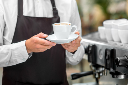 Barista in the brown apron holding white coffee cup in the cafeの写真素材