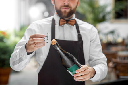 Handsome barman in uniform opening bottle with sparkling wine outdoor on the restaurant terraceの写真素材