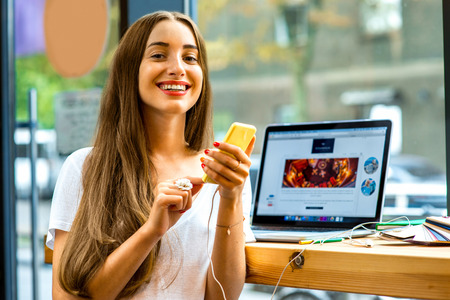 Young woman using yellow smart phone sitting near the window with laptop and colorful stuff on the table in the cafeの写真素材