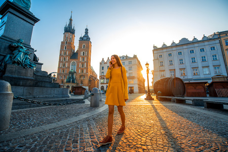 Young and beautiful woman in yellow dress walking and talking with mobile phone in the old city center of Krakow on the morning sunriseの写真素材