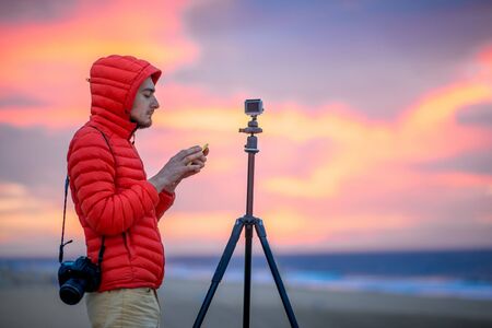 Photographer in red jacket with hood filming with small action camera on the tripod beautiful sunrise on the sand dunes in the early morning.の写真素材