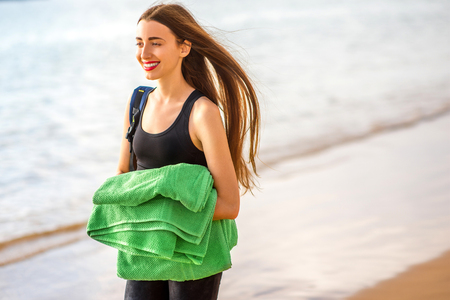 Young woman in black sport clothes walking with green towel and backpack on the beachの写真素材