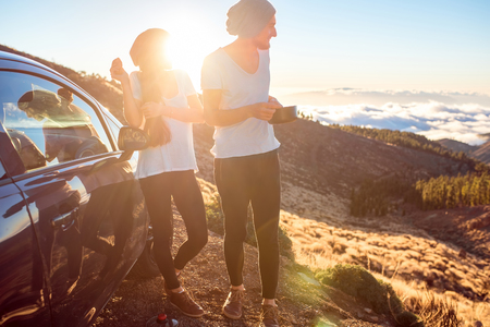Young couple in white t-shirts and hats having picnic near the car on the mountain roadside above the cloudsの写真素材