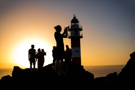 Silhouettes of people on the rocky coastline near the lighthouse. Deep contrasted yellow and black imageの写真素材