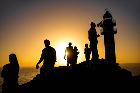 Silhouettes of people on the rocky coastline near the lighthouse. Deep contrasted yellow and black imageの写真素材