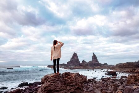 Young woman in sweater standing on the rocky coast and looking forward on the cliffs on the cloudy weather. Wide angle image, rear view with a lot of spaceの写真素材