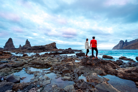 Young and romantic couple in red sweaters standing together and looking forward on the rocky coast on the cloudy weather. Wide angle image, rear view with copy spaceの写真素材