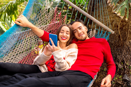 Young couple in sweaters using smart phone sitting together on the hammock in the gardenの写真素材