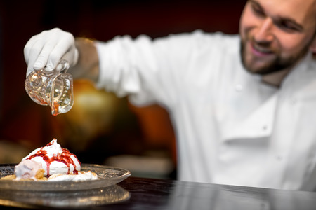 Chef cook pouring jam on the pavlova dessert on the bar table at the open kitchen of the restaurant. Close-up image with small depth of field focused on the dessertの写真素材