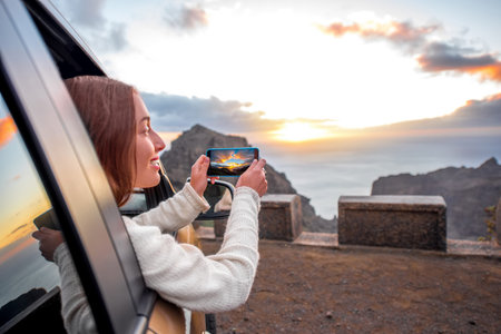Young female traveler photographing with smartphone beautiful rocky coast on the sunset.