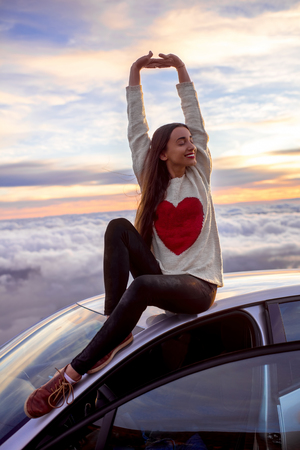 Young woman in sweater with heart shape rising hands sitting on the car roof above the clouds on the sunrise.の写真素材