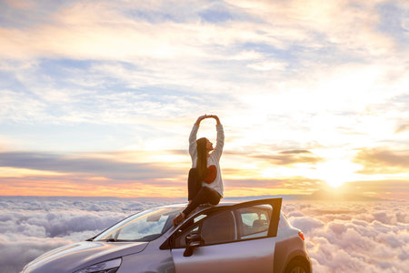 Young woman in sweater with heart shape rising hands sitting on the car roof above the clouds on the...