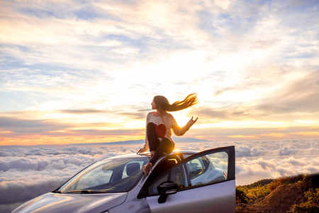 Young woman in sweater with heart shape enjoying beautiful cloudscape sitting on the car roof above the clouds on the sunrise.の写真素材