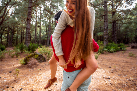 Young playful couple in sweaters and hats having a piggyback ride in the pine forest. Strong cropped image compositionの写真素材