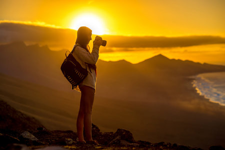 Young female traveler with backpack and photo camera photographing beautiful Cofete coastline with...