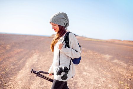 Young female photographer in sweater and hat walking with camera, backpack and tripod on the...