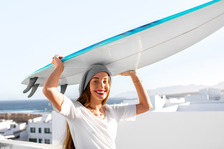 Portrait of a young smiling woman dressed casual carrying surfboard on the white city background.の写真素材