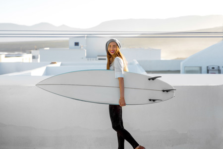 Young smiling woman in white t-shirt and hat standing with surfboard on the roof top on the white city background.の写真素材