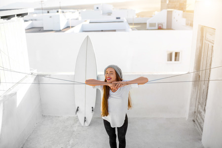 Young smiling woman in white t-shirt and hat standing on the roof top with surfboard and white buildings on the background in the morningの写真素材