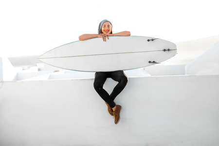 Young smiling woman in white t-shirt and hat sitting with surfboard on the roof top on the white city background.の写真素材