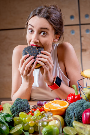 Hungry woman eating black burger at the table full of fruits and vegetables on the wooden background. Choosing between healthy and unhealthy foodの写真素材