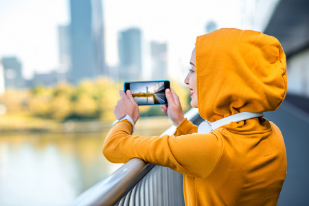 Young woman in yellow hood photographing modern city on the  bridge with skyscrapers on the background in the morningの写真素材