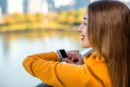 Young woman in yellow sweater using smart watch on the modern bridge with skyscrapers on the background. Sport application on the smart watchの写真素材
