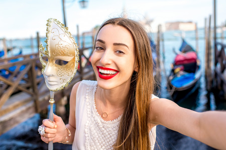 Young female traveler making selfie photo with carnaval mask standing near San Marco square with gondolas on the background in Venice.の写真素材