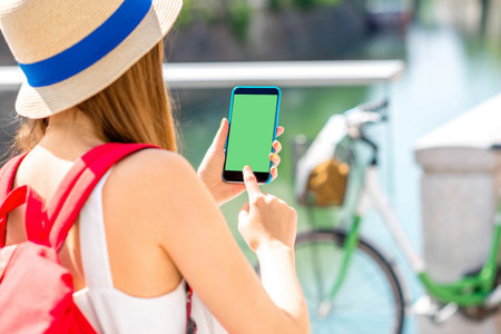 Female traveler holding smart phone with green screen on the street with bicycle on the background.