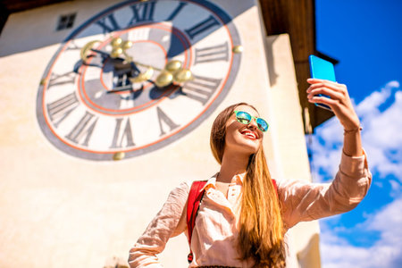 Young female traveler making selfie photo with smart phone on the old clock tower background in Graz...